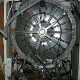 Wnętrze pralki z widocznym bębnem, paskiem napędowym i okablowaniem, podczas naprawy, z kluczem nasadowym opartym o obudowę.