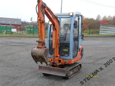 Pomarańczowo-niebieska koparka na gąsienicach, z łyżką skierowaną do przodu, stojąca na szarym placu. Widoczne budynki w tle.