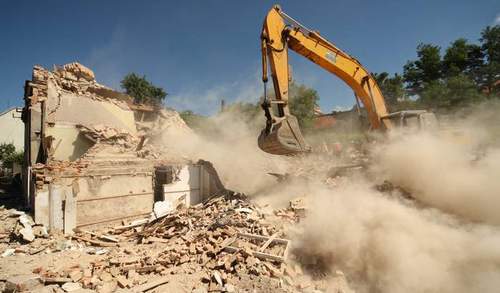 Żółta koparka burzy fragment budynku w obłokach kurzu i gruzu.