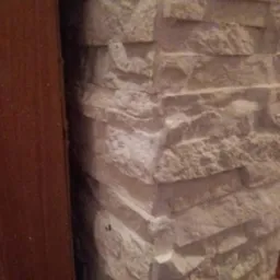 Dekoracyjna ściana wykończona jasnym kamieniem elewacyjnym ułożonym warstwowo, przylegająca do drewnianej framugi drzwi.