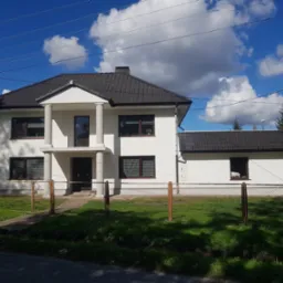 Dwukondygnacyjny dom z białą elewacją, ciemną dachówką i kolumnami przy wejściu, otoczony zielonym trawnikiem i ogrodzeniem z siatki.