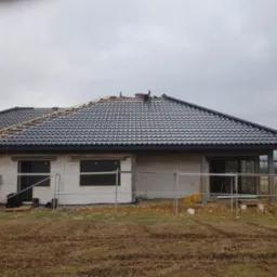 Dom w trakcie budowy z częściowo ułożonym ciemnoszarym dachem i widoczną więźbą dachową, rusztowania przy ścianach z betonu komórkowego, pochmurne niebo.