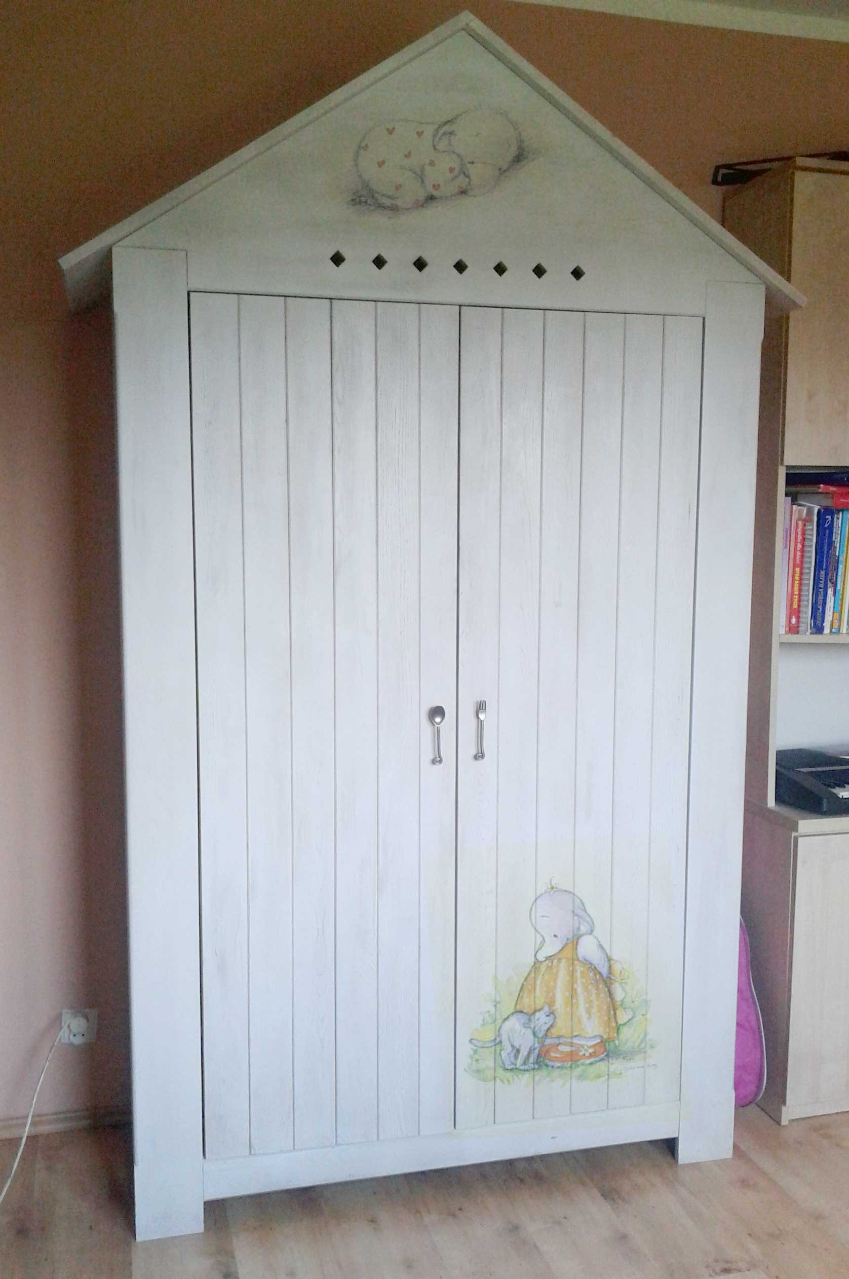 Biała, drewniana szafa dziecięca z motywem śpiącego słonia na szczycie i rysunkiem dziecka z kotem na drzwiach, uchwyty w kształcie sztućców, widoczne elementy pokoju dziecięcego.