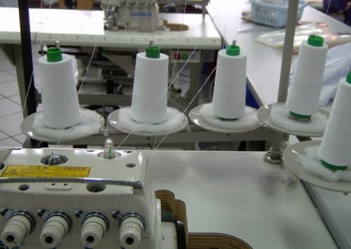 Fragment maszyny szwalniczej z nawiniętymi białymi nićmi na szpulach z zielonymi zakończeniami, widoczne naprężacze nici i żółta naklejka ostrzegawcza.