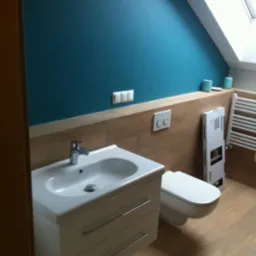 Turkusowa ściana w łazience ze skośnym sufitem, biała umywalka z szafką, wisząca toaleta i grzejnik drabinkowy na tle płytek imitujących drewno.