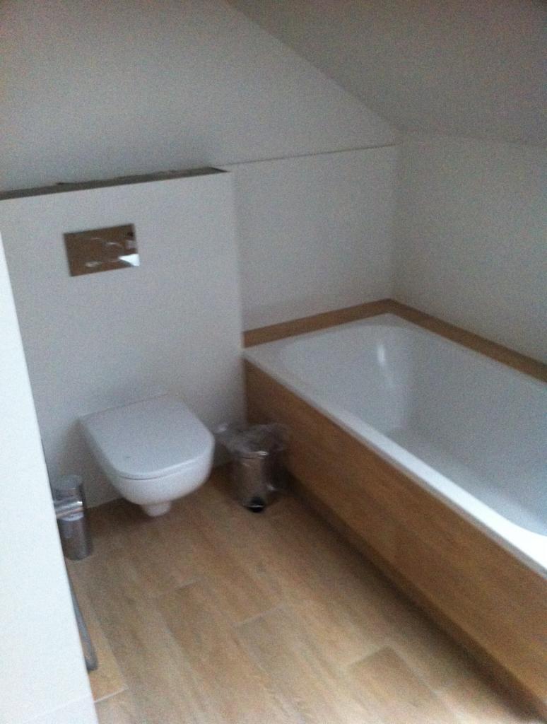 Nowoczesna łazienka na poddaszu z drewnopodobnymi płytkami podłogowymi, zabudowaną toaletą i wanną obudowaną drewnem.