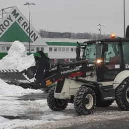 Biała koparko-ładowarka marki Hidromek z pełną łyżką śniegu na parkingu sklepu Leroy Merlin, światła ostrzegawcze włączone.