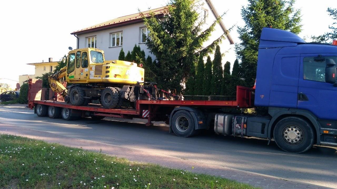 Niebieska ciężarówka z niskopodwoziową naczepą przewożąca żółtą koparkę kołową; w tle dom jednorodzinny i iglaki.
