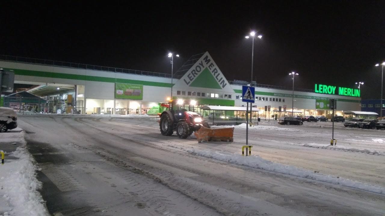 Czerwony traktor z pługiem odśnieża oświetlony parking przed budynkiem Leroy Merlin w zimowej scenerii nocą.