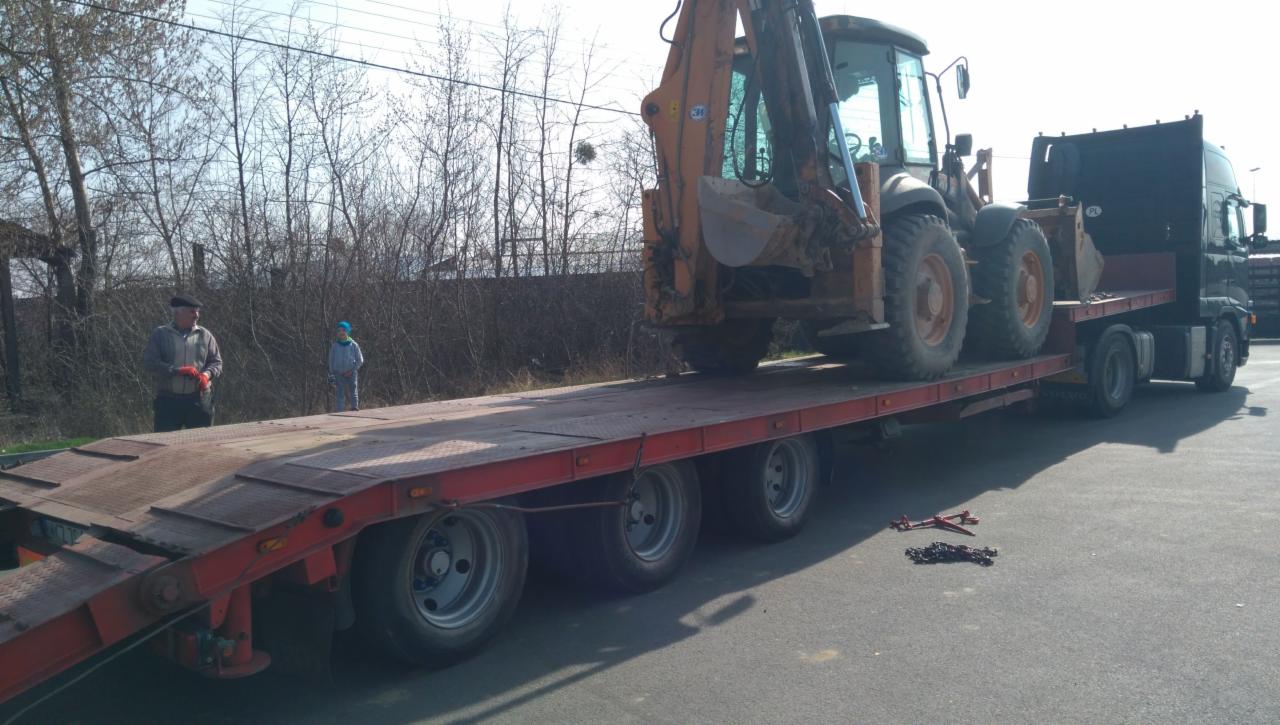 Pomarańczowa koparko-ładowarka transportowana na niskopodwoziowej lawecie, zabezpieczona łańcuchami, z dwoma osobami obserwującymi na rampie w tle.
