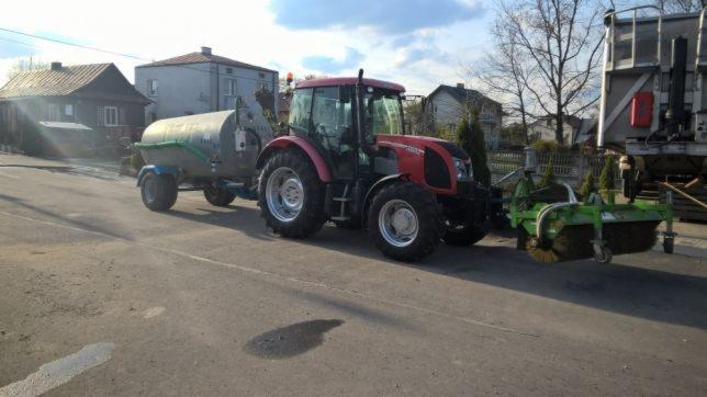 Czerwony traktor z przyczepą cysterną i szczotką czyszczącą jezdnię na tle wiejskich domów w słoneczny dzień.