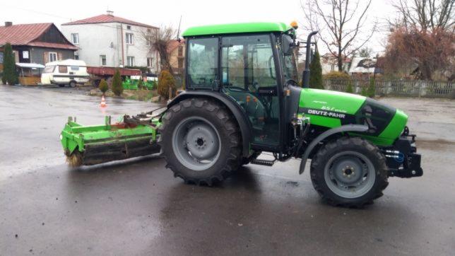 Zielony traktor Deutz-Fahr z zamontowaną szczotką do zamiatania na mokrym, asfaltowym placu, w tle budynki mieszkalne i przyczepa kempingowa.