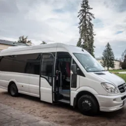 Biały bus Mercedes-Benz Sprinter z otwartymi drzwiami pasażera, zaparkowany na żwirowej nawierzchni przed klasycystycznym budynkiem z fontanną w tle.