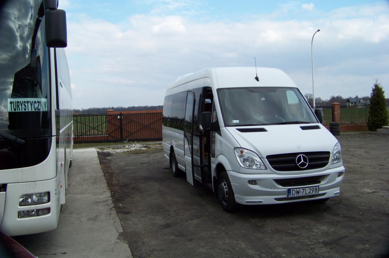 Biały bus Mercedes-Benz z otwartymi drzwiami, zaparkowany obok autokaru z napisem 'Turystyczny', na tle ogrodzenia i pochmurnego nieba.