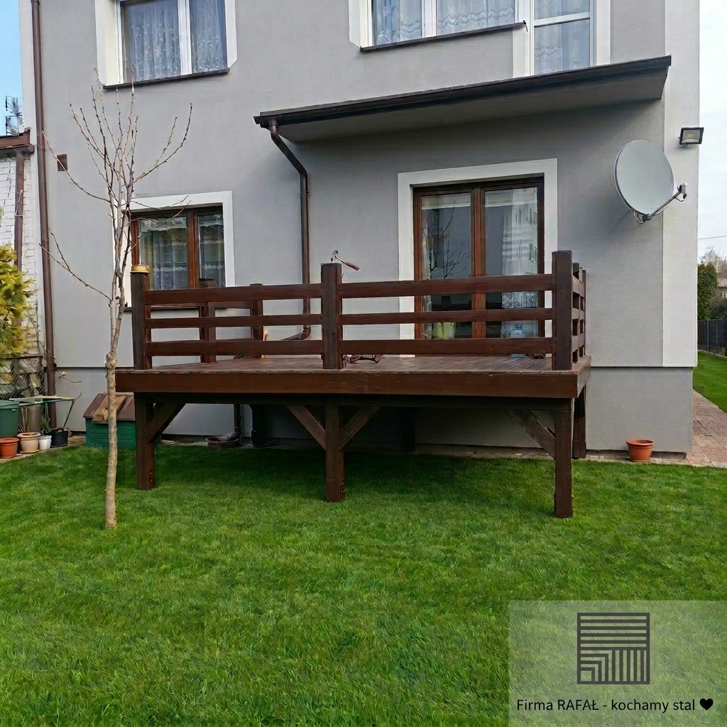 Drewniany taras z balustradą przy szarym domu, widok z poziomu trawnika. Ciemne drewno kontrastuje z zielenią trawy i jasną elewacją. Widoczne okna i satelita na ścianie.