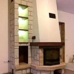Nowoczesny kominek z obudową z jasnego kamienia, z wnękami oświetlonymi od wewnątrz, ustawiony na brązowych płytkach podłogowych, widoczne gniazdko elektryczne na ścianie.