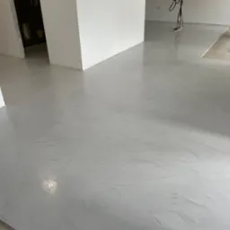 Jasnoszara posadzka z efektem mikrocementu, widoczne delikatne wzory i refleksy światła, fragment pomieszczenia w trakcie prac wykończeniowych, widoczne puszki z farbą i narzędzia w tle.