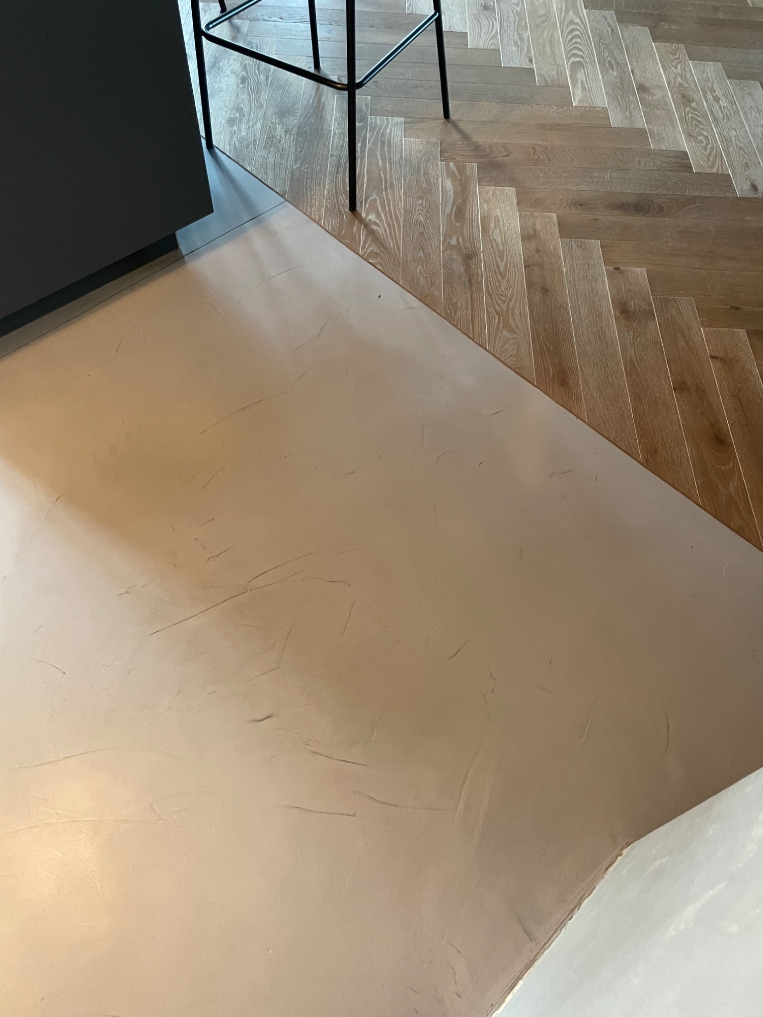 Jasna posadzka mikrocementowa z widoczną fakturą, kontrastująca z drewnianą podłogą ułożoną w jodełkę i fragmentem czarnego stołka barowego.