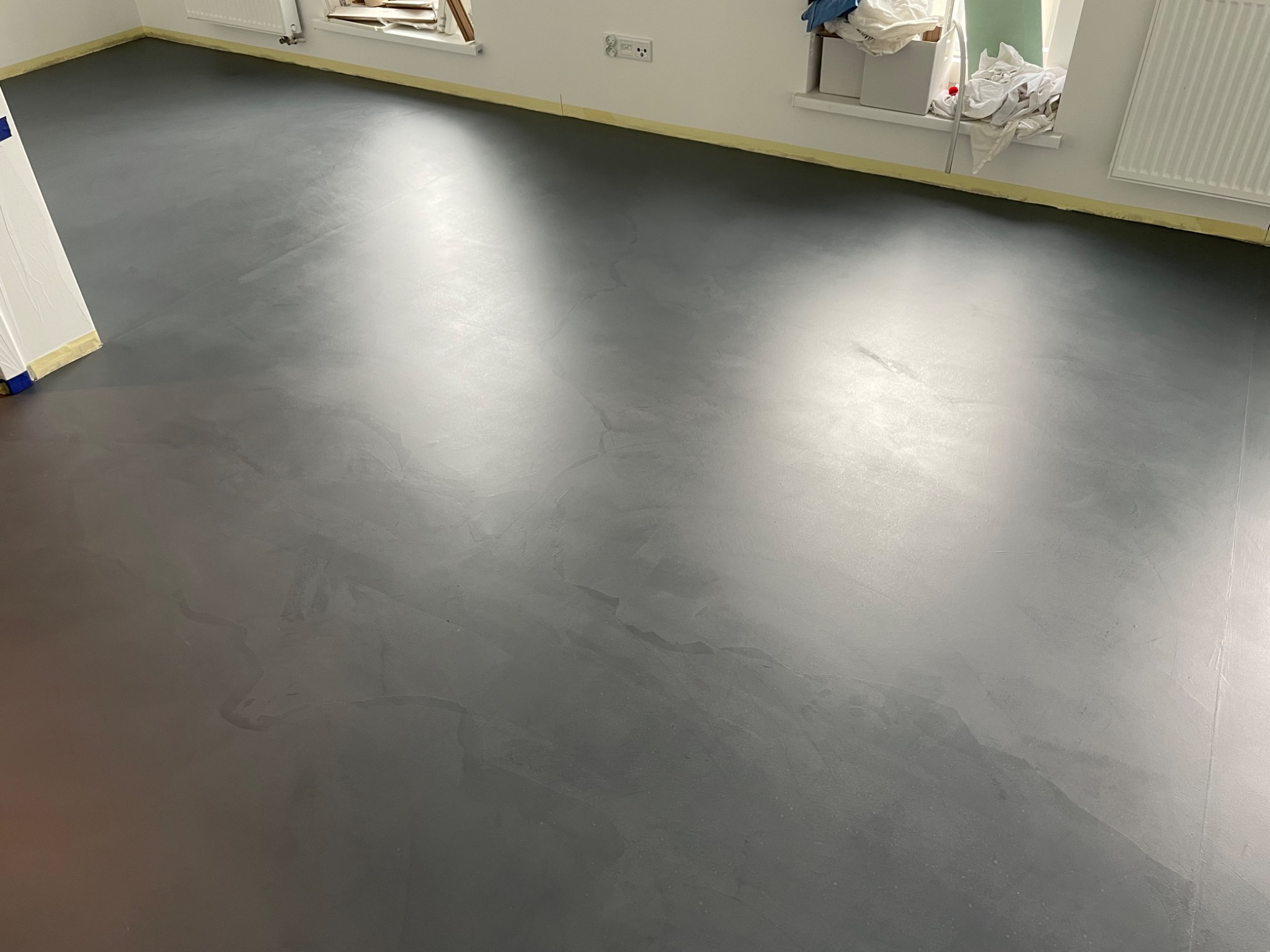 Szara, gładka posadzka z mikrocementu w jasnym pomieszczeniu, zabezpieczona taśmą malarską przy ścianach i drzwiach.