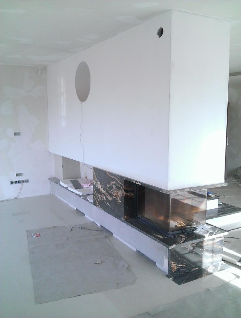 Nowoczesny kominek z czarnym marmurem w Myślenicach. Biała obudowa z okrągłym otworem wentylacyjnym. Widoczne elementy montażowe i zabezpieczenia podłogi.