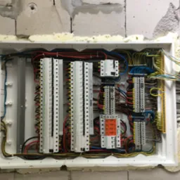 Rozdzielnica elektryczna w trakcie montażu, widoczne kable, bezpieczniki i listwy zaciskowe, osadzona w ścianie z betonu komórkowego, uszczelniona pianką montażową.
