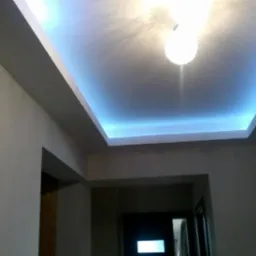 Podwieszany sufit z oświetleniem LED w kolorze niebieskim, centralnie umieszczona lampa sufitowa, widok z dołu na korytarz z drzwiami w tle.