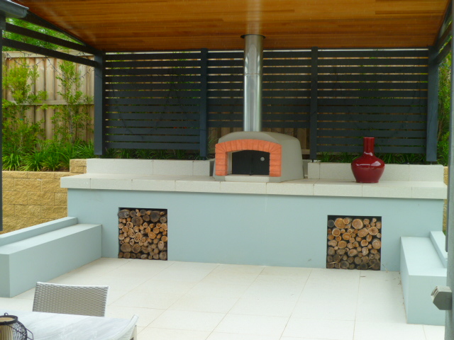 Zadaszony, murowany piec do pizzy z pomarańczową obudową paleniska i zapasem drewna w niszach, uzupełniony czerwoną wazą na blacie, otoczony jasną, wyłożoną płytkami przestrzenią.