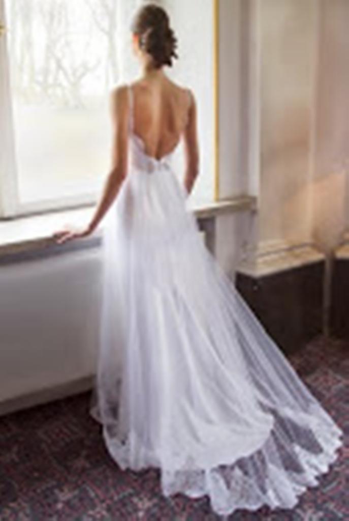 Kobieta w białej, zwiewnej sukni ślubnej z odkrytymi plecami, stojąca tyłem przy oknie z widokiem na drzewa. Suknia ma delikatne ramiączka i długi tren.