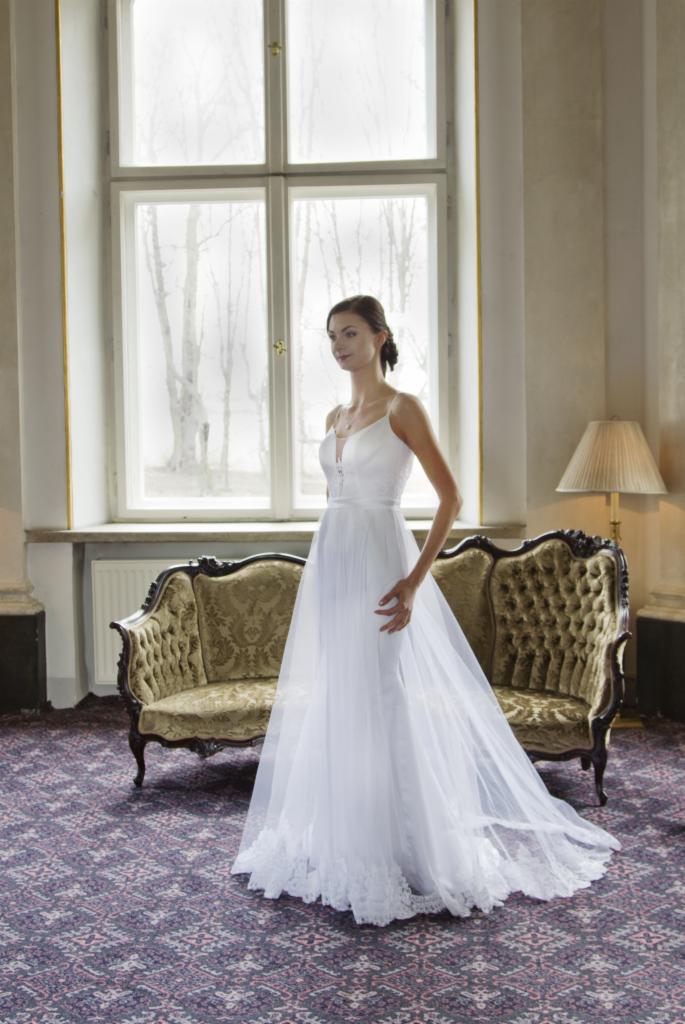 Elegancka kobieta w białej sukni ślubnej z delikatnymi ramiączkami, pozuje na tle okna i sofy w klasycznym wnętrzu. Długa, zwiewna suknia z koronkowym wykończeniem.