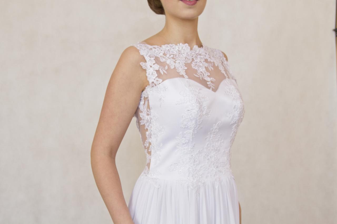 Elegancka suknia ślubna z koronkową górą i delikatnym, zwiewnym dołem, prezentowana na modelce w neutralnym otoczeniu.