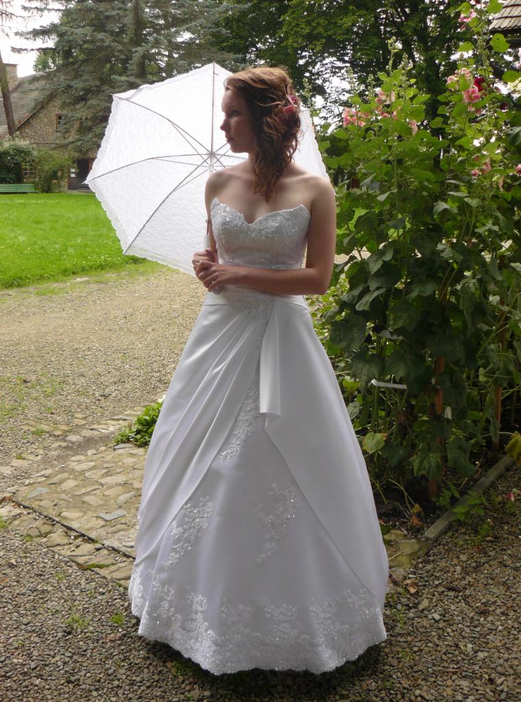 Panna młoda w białej sukni ślubnej z parasolem, ogród w tle. Szycie odzieży na miarę w Krakowie. Delikatne zdobienia i romantyczna stylizacja.