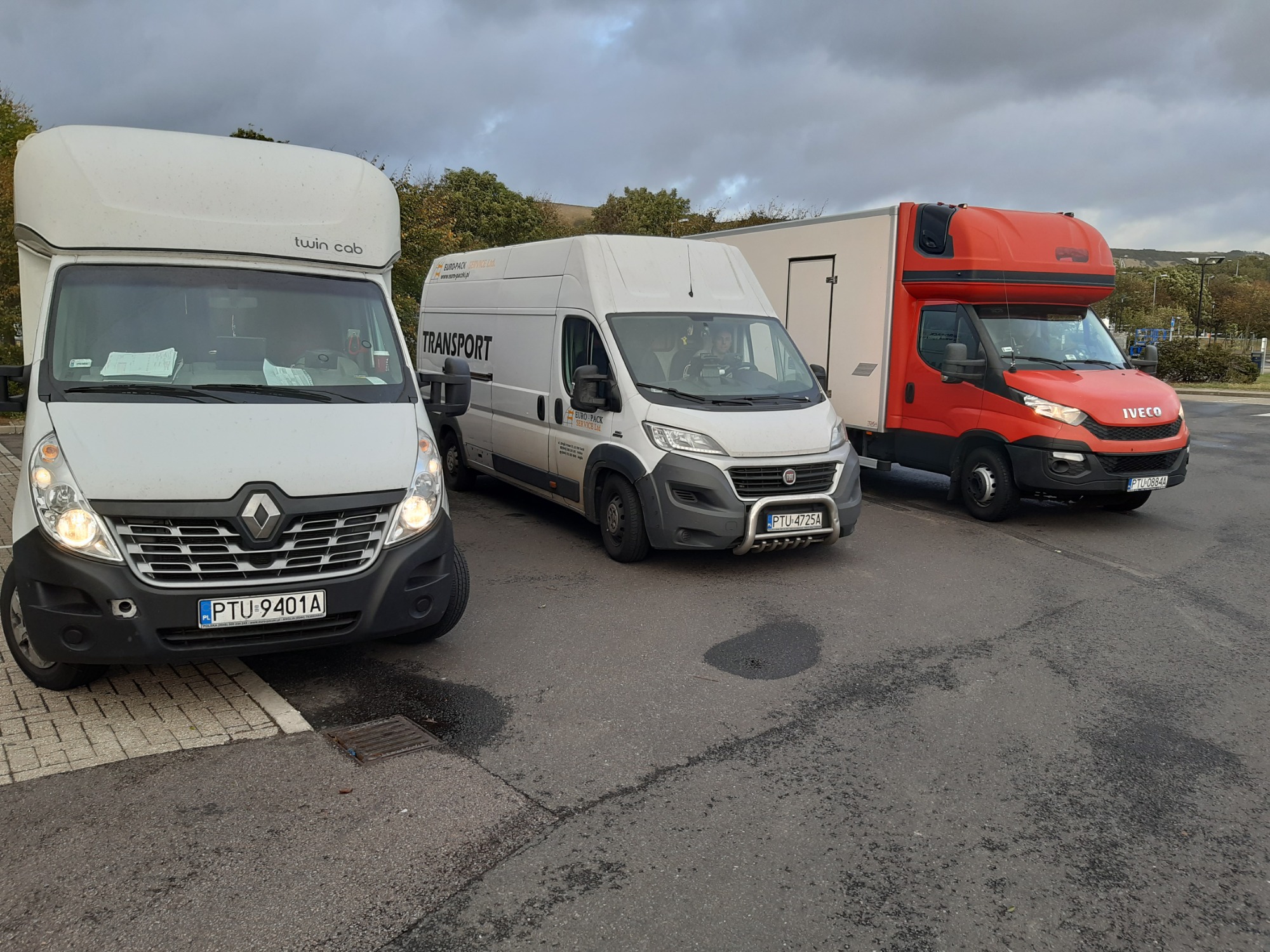 Trzy busy transportowe różnych marek (Renault Master, Fiat Ducato, Iveco) zaparkowane obok siebie na parkingu, z widocznymi numerami rejestracyjnymi i oznaczeniami firm transportowych.