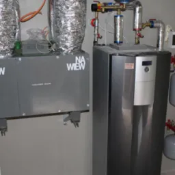 Szara instalacja grzewcza z dwoma srebrnymi rurami wentylacyjnymi, zbiornikami wyrównawczymi i licznymi zaworami w pomieszczeniu o szarych ścianach.