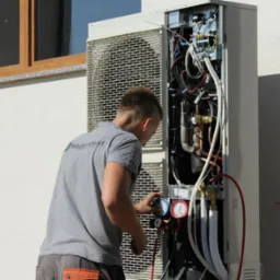 Mężczyzna w szarej koszulce z logo firmy sprawdza ciśnienie w otwartej obudowie pompy ciepła na tle elewacji budynku z oknem.