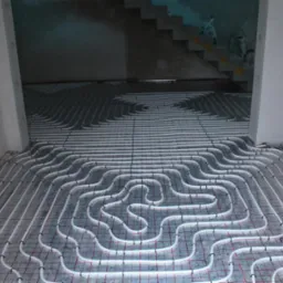 Białe rury ogrzewania podłogowego ułożone na siatce w surowym wnętrzu, widoczne schody w tle.