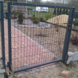 Otwarta furtka z metalowej siatki, prowadząca na ścieżkę z czerwonej kostki brukowej, widoczny ogród w trakcie aranżacji, w tle przyczepa kempingowa.