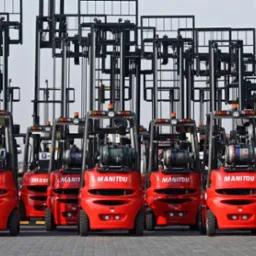 Rząd czerwonych wózków widłowych Manitou na placu, z podniesionymi masztami i butlami gazowymi, ustawionych równo w rzędach, widok z przodu, oświetlenie dzienne.