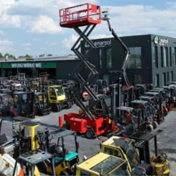 Szeroki kadr na plac z wózkami widłowymi i podnośnikiem nożycowym Lemarpol, budynek firmy w tle, słoneczny dzień.