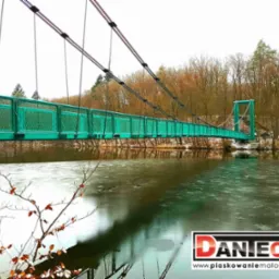 Zielony most wiszący nad zamarzniętym jeziorem, widok z dołu, w tle drzewa bez liści i logo firmy Daniech w prawym dolnym rogu.