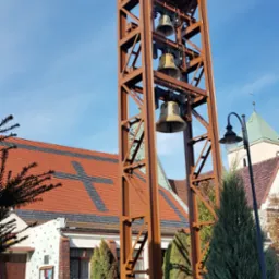 Stalowa konstrukcja dzwonnicy z trzema dzwonami na tle kościoła z dachem z krzyżem i błękitnego nieba.