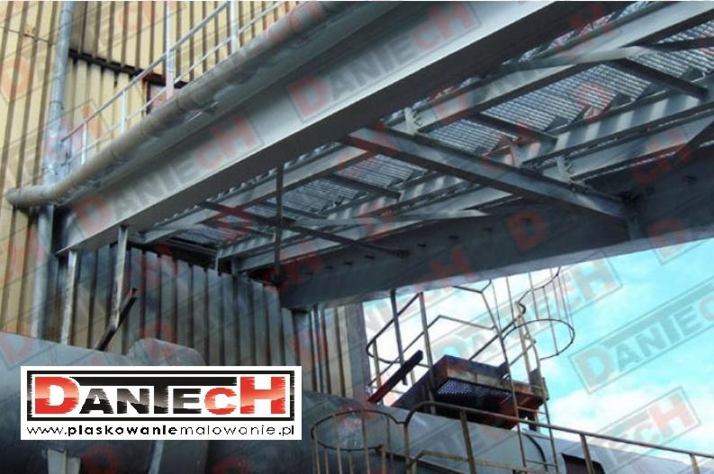 Konstrukcja stalowa pomostu technologicznego z krat pomostowych i rur, wsparta na elementach budynku przemysłowego, widok od dołu, z logo firmy w lewym dolnym rogu.