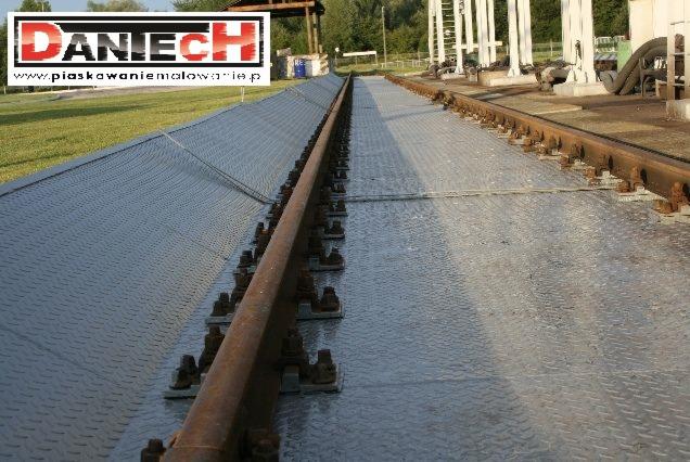 Szyny kolejowe na nowej, pomalowanej proszkowo platformie z blachy ryflowanej, widoczne ślady rdzy na szynach i śrubach mocujących.