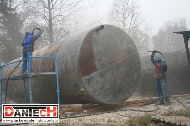 Dwaj pracownicy w kombinezonach ochronnych, z użyciem specjalistycznego sprzętu, czyszczą duży, metalowy zbiornik na zewnątrz, otoczeni chmurą pyłu.