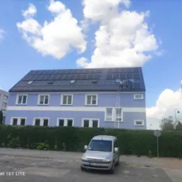 Dwóch monterów instaluje panele słoneczne na dachu trzypiętrowego budynku mieszkalnego o niebieskiej fasadzie, drabina oparta o ścianę, na pierwszym planie zaparkowany srebrny samochód dostawczy.