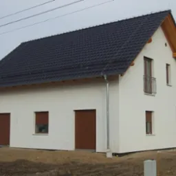 Biały dom z ciemną dachówką i brązowymi roletami zewnętrznymi. Widoczny fragment balkonu i elementy drewniane w konstrukcji dachu. Surowy teren wokół domu.