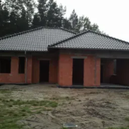 Surowy dom z czerwonej cegły z ciemną dachówką na tle lasu. Widoczne otwory okienne i drzwiowe. Stan surowy zamknięty z wykonanym dachem.