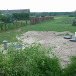 Instalacja przydomowej oczyszczalni ścieków z dwoma włazami i skrzynką sterującą na piaszczystym podłożu, widok zza krzewów, w tle ogrodzenie i łąka.