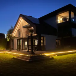 Nowoczesny dom jednorodzinny z oświetloną elewacją i tarasem w scenerii nocnej, widoczne palmy i podświetlony trawnik.