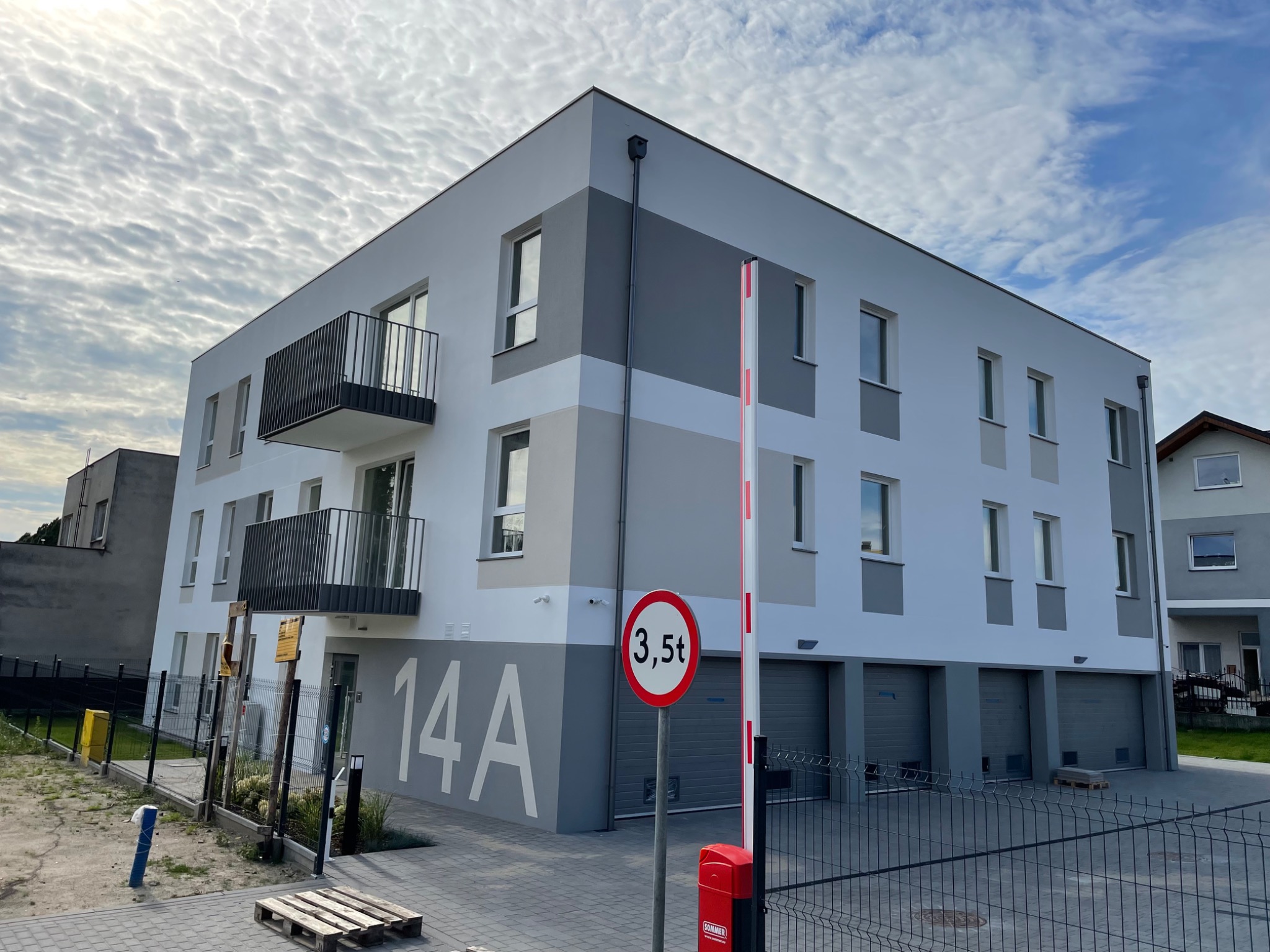 Nowoczesny, trzypiętrowy budynek mieszkalny z balkonami i garażami, pomalowany w odcieniach bieli i szarości, z numerem 14A na elewacji, znak drogowy ograniczający tonaż i szlaban na pierwszym...
