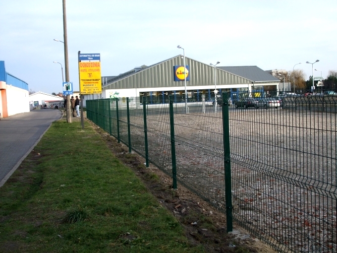 Zielona siatka ogrodzeniowa 3D zabezpieczająca parking przed sklepem Lidl, widok z zewnątrz, słupki osadzone w ziemi, w tle zaparkowane samochody i budynek sklepu.